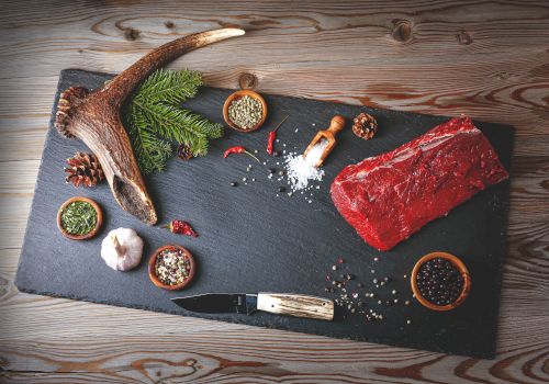 A slate board with a raw red steak, pine branches, an antler, spices, salt, pepper, and a knife on a rustic wooden table, ready for grilling.