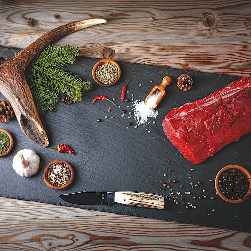 A slate board with a raw red steak, pine branches, an antler, spices, salt, pepper, and a knife on a rustic wooden table, ready for grilling.