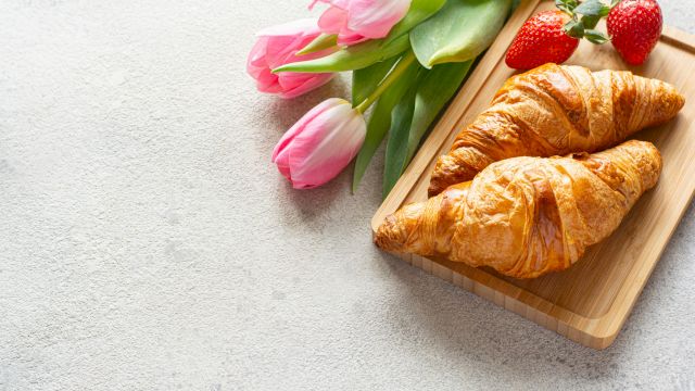 Two croissants on a wooden tray, accompanied by pink tulips and strawberries, placed on a light textured surface.