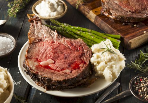 A slice of juicy medium-rare steak with bone, creamy mashed potatoes, asparagus, herbs, and peppery sides on a rustic board.