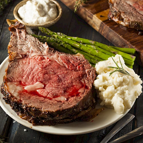 A slice of juicy medium-rare steak with bone, creamy mashed potatoes, asparagus, herbs, and peppery sides on a rustic board.