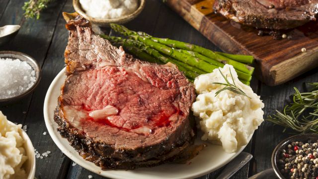 A slice of juicy medium-rare steak with bone, creamy mashed potatoes, asparagus, herbs, and peppery sides on a rustic board.