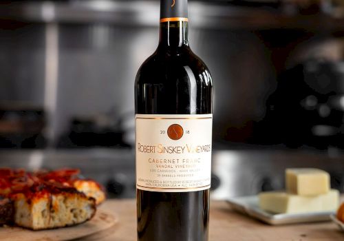 A bottle of red wine on a wooden table with a pizza slice and butter in the background, all set in a cozy kitchen scene.
