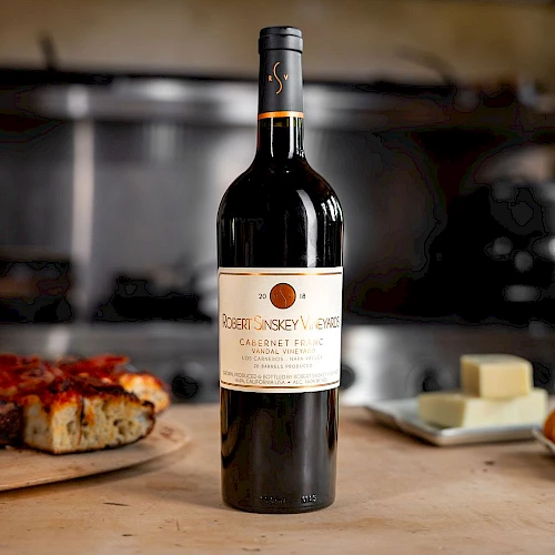 A bottle of red wine on a wooden table with a pizza slice and butter in the background, all set in a cozy kitchen scene.