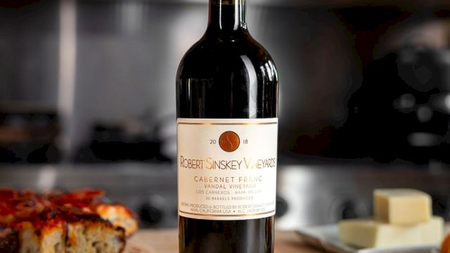 A bottle of red wine on a wooden table with a pizza slice and butter in the background, all set in a cozy kitchen scene.