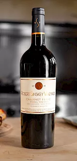 A bottle of Cabernet Franc wine on a table with bread and cheese in a softly lit background.