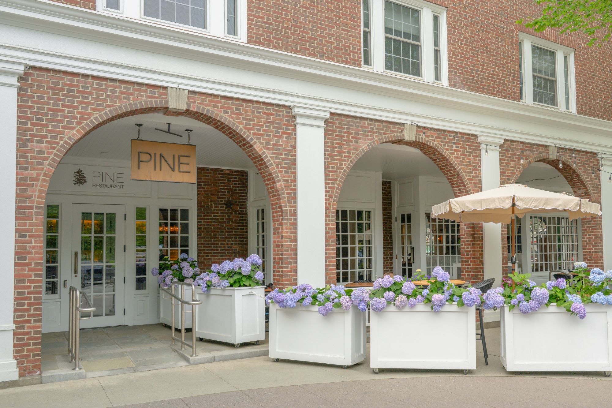 Brick entrance of Pine Restaurant with arched doorways, planters of purple hydrangeas, and an outdoor patio umbrella nearby.