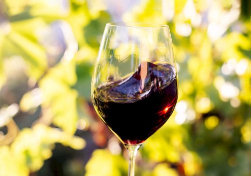 A glass of red wine sits in soft-focus outdoor light, with a warm, celebratory vibe in the background.