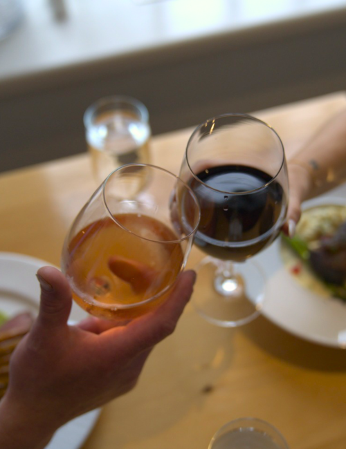 Two people clink glasses of red and amber wine at a table, toasting together.