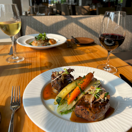 A restaurant table with steak, roasted carrots, and microgreens, plus two glasses of wine in bright sunlight.