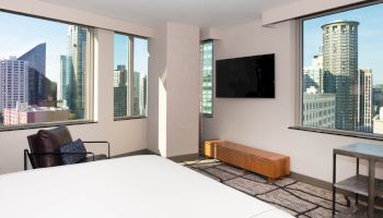 The image shows a modern hotel room with city views, featuring large windows, a bed, TV, and minimalist furniture.