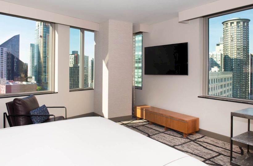 The image shows a modern hotel room with city views, featuring large windows, a bed, TV, and minimalist furniture.