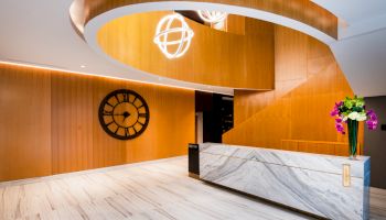 The image shows a modern lobby with a marble reception desk, a large wall clock, wooden paneling, and decorative lighting fixtures.
