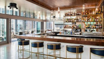 A modern bar with blue stools, a wooden counter, and shelves stocked with bottles against a backdrop of large windows and urban scenery.