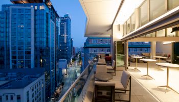A modern balcony overlooks a cityscape with tall buildings at dusk, featuring outdoor seating and tables under soft lighting.