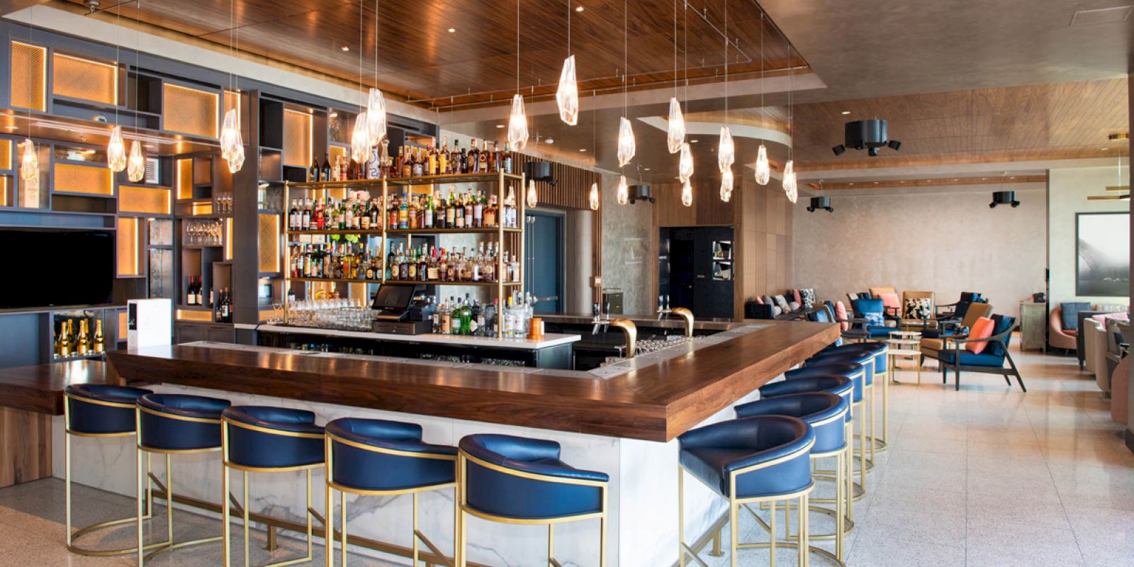 This image shows a modern, stylish bar with wooden accents, pendant lights, and blue bar stools arranged around the corner of the bar area.