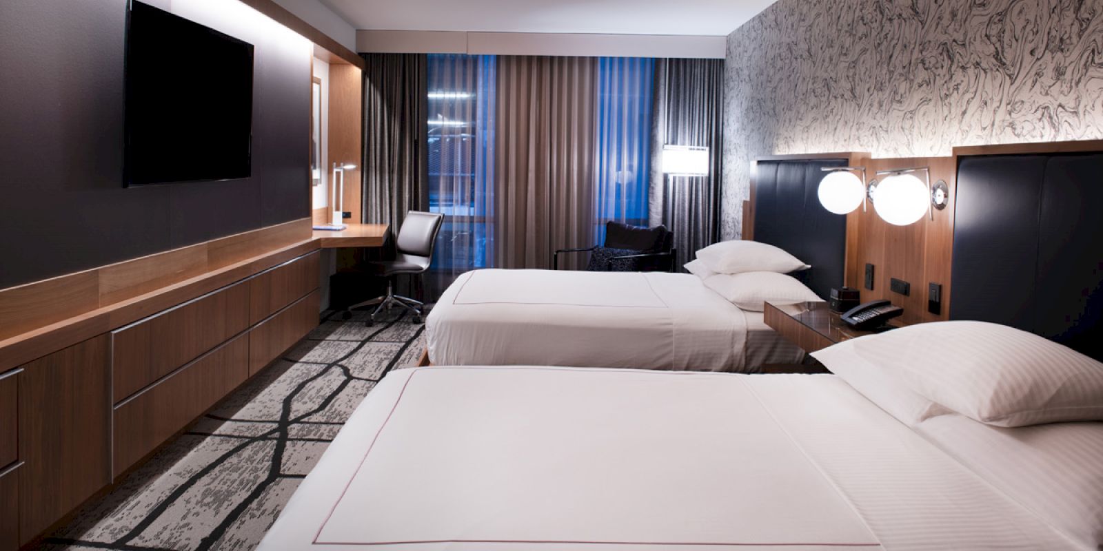 A modern hotel room with two beds, a wall-mounted TV, desk, and a window with curtains. Stylish decor and clean lines complete the look.