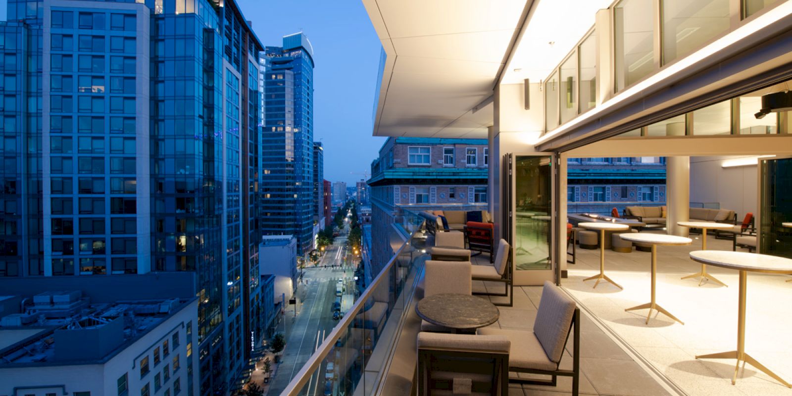 A modern cityscape with high-rise buildings at dusk, featuring a rooftop terrace with tables and chairs overlooking the street below.