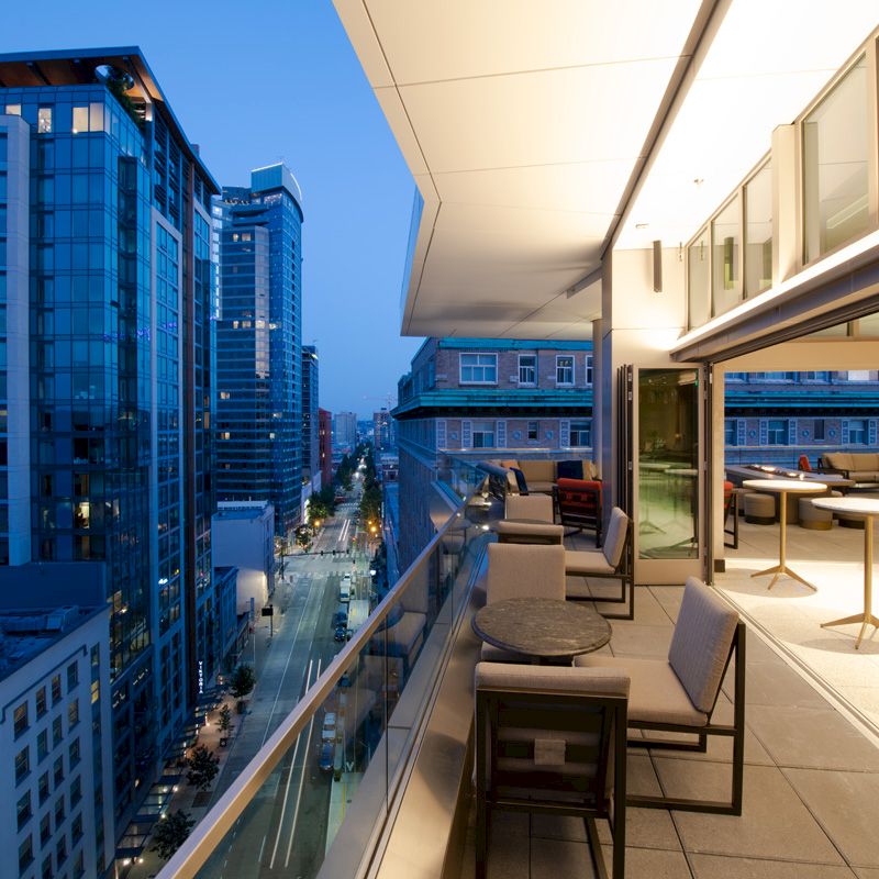 A modern rooftop patio with seating overlooks a cityscape with tall buildings at dusk, featuring a blend of outdoor and indoor spaces.