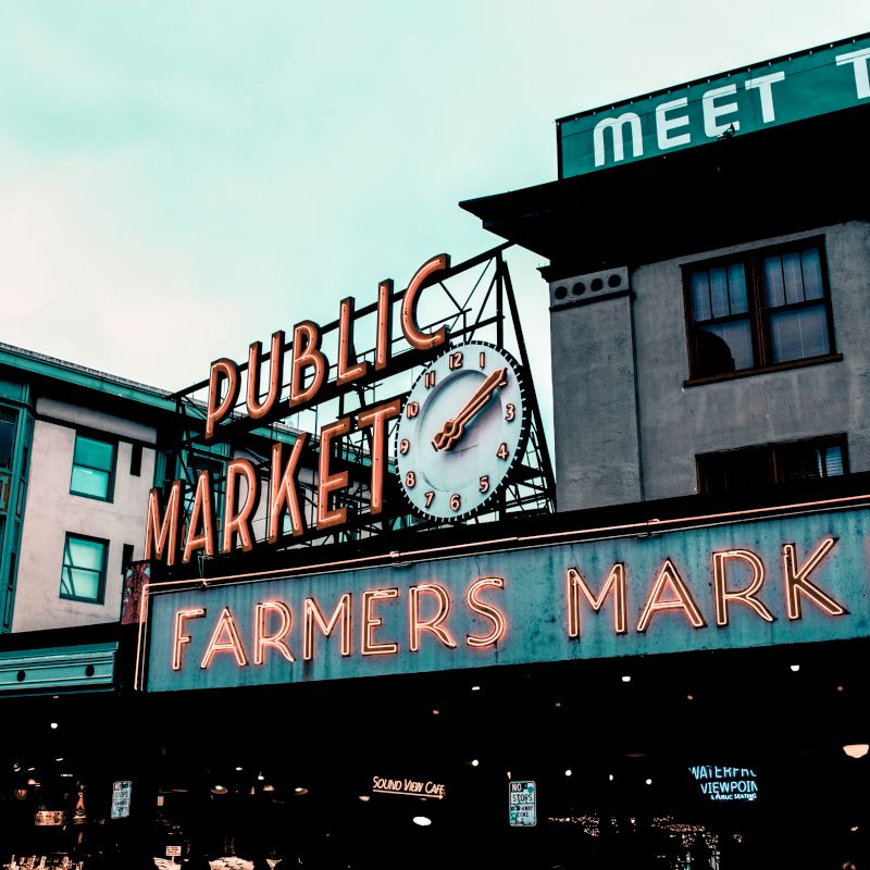 The image shows a neon sign for a "Public Market" with a clock and "Farmers Market," set against urban buildings and an overcast sky.