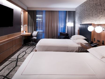 A modern hotel room with two beds, a flat-screen TV, a desk with a chair, and stylish lighting and decor.