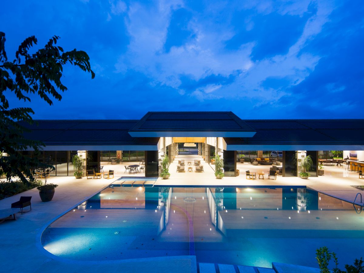 The image shows a luxurious outdoor swimming pool at night with a modern building and open-air seating in the background.