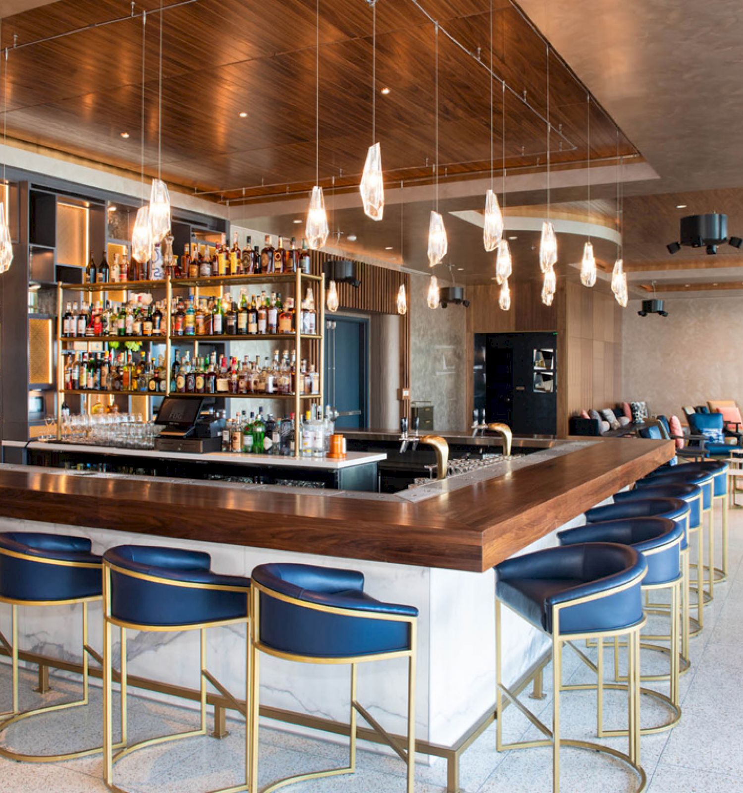 A modern bar with a wooden counter and blue seating, surrounded by shelves of bottles and pendant lights, in a spacious room.