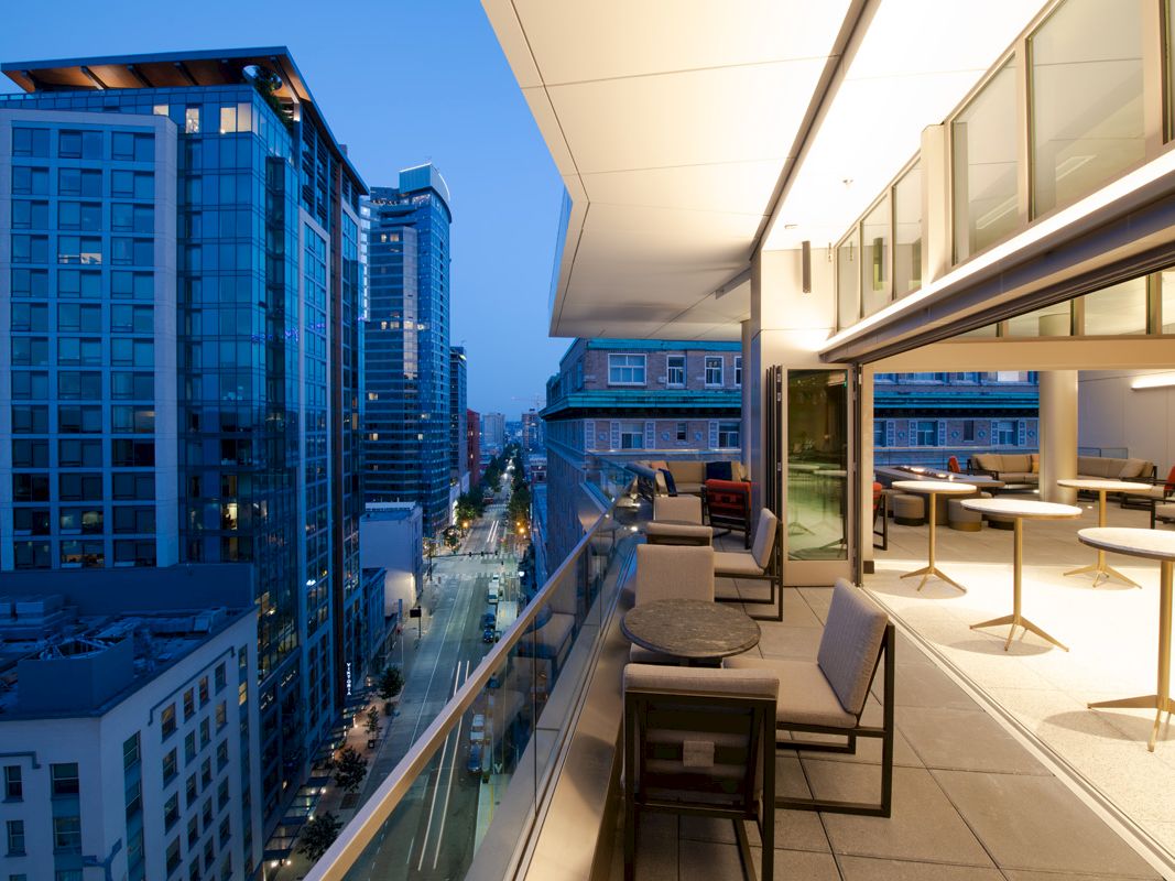 A modern rooftop terrace with tables and chairs overlooks a cityscape, surrounded by tall buildings under a clear evening sky.