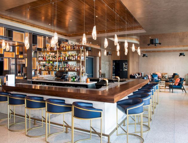 A modern bar interior with a central counter, stylish bar stools, a variety of bottles, and hanging pendant lights. Cozy seating area visible.