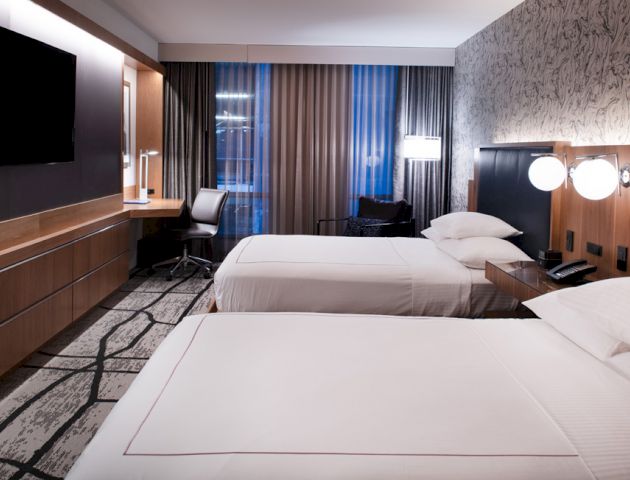 A modern hotel room features two beds, a TV, a work desk, stylish decor, and large windows with curtains, creating a comfortable atmosphere.