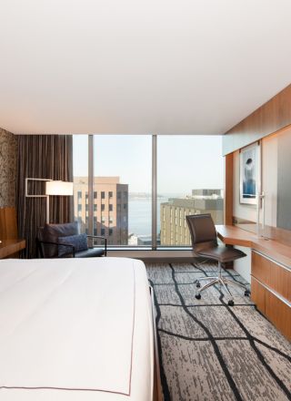 A modern hotel room with a large bed, desk, chair, TV, and a city view through a wide window. The decor is contemporary and stylish.