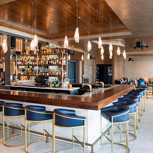 A modern bar with a well-stocked shelf, blue chairs, and pendant lights. The space is open and stylish, featuring a lounge area.