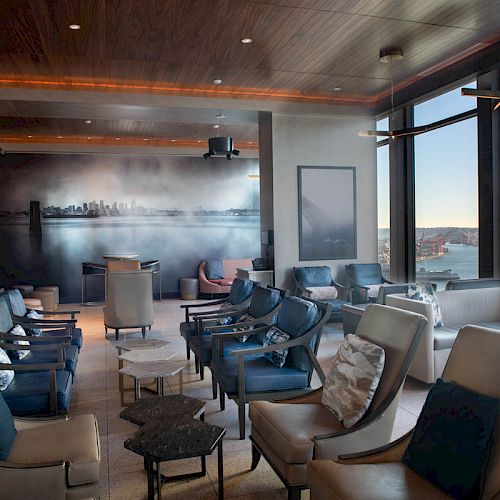 The image shows a modern lounge with comfortable seating, large windows offering a scenic view, and a mural of a city skyline on the wall.