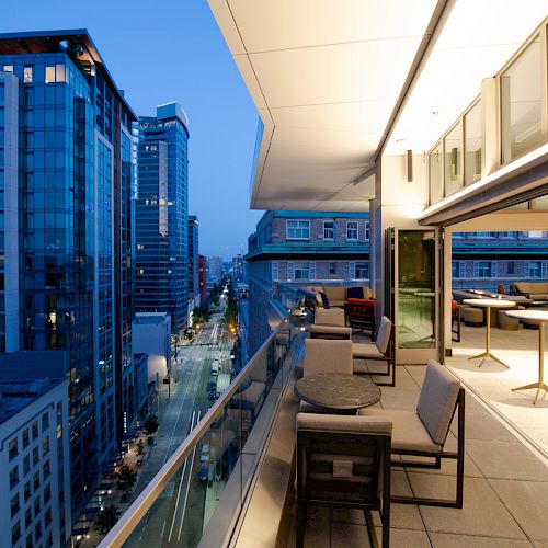 A balcony with tables and chairs overlooks a city street lined with tall buildings during evening.