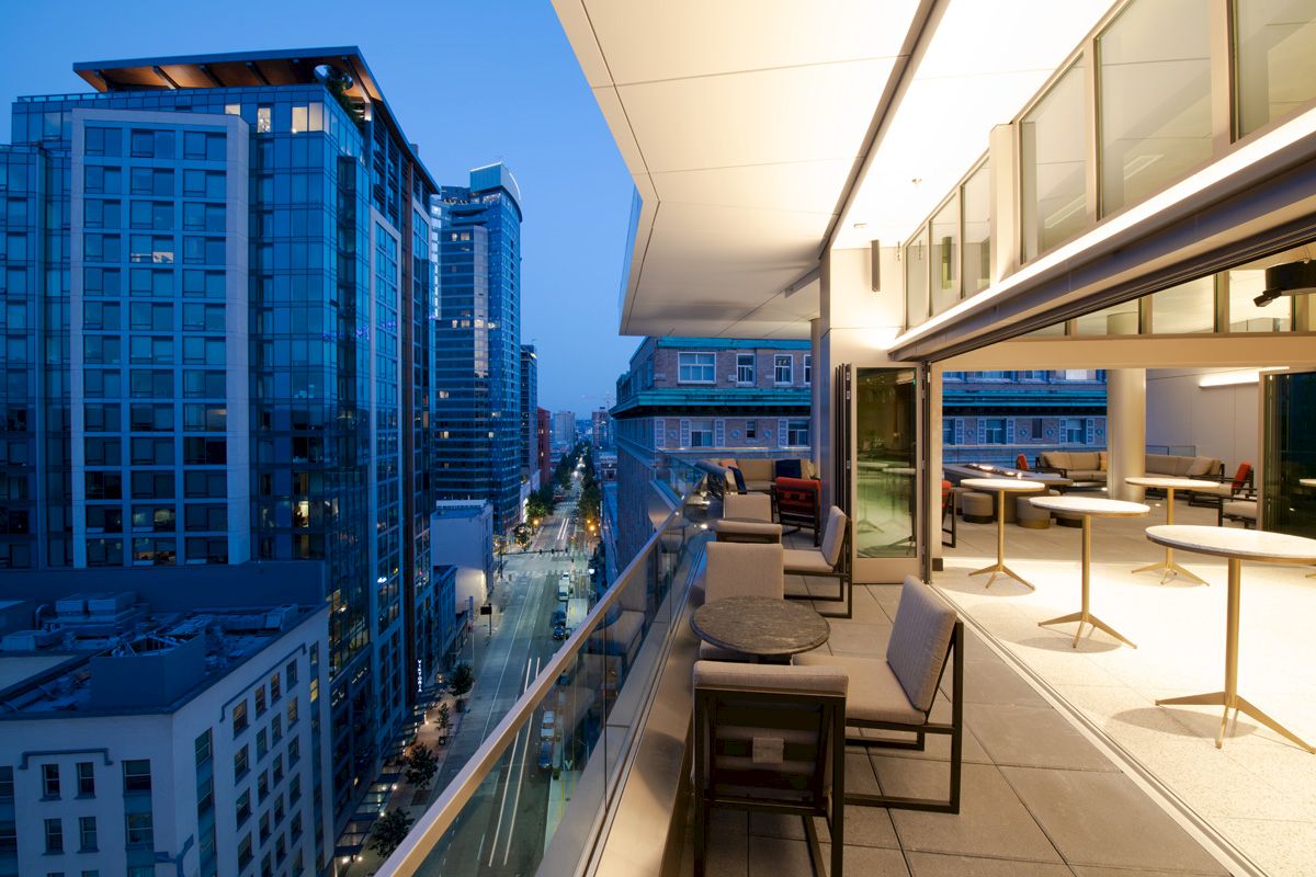 A modern outdoor terrace with seating overlooks a cityscape of tall buildings and streets at dusk, creating an inviting urban atmosphere.