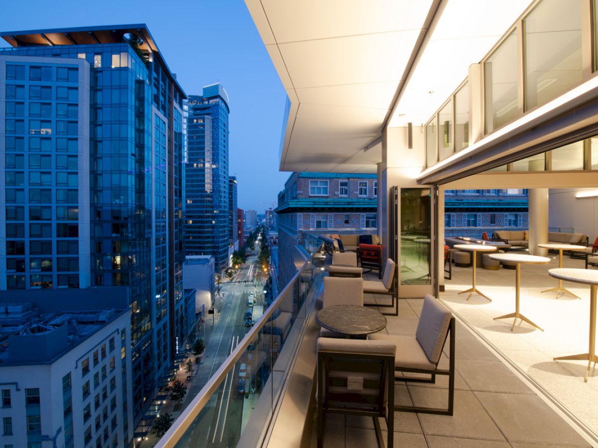 A modern outdoor terrace with seating overlooks a cityscape of tall buildings and streets at dusk, creating an inviting urban atmosphere.