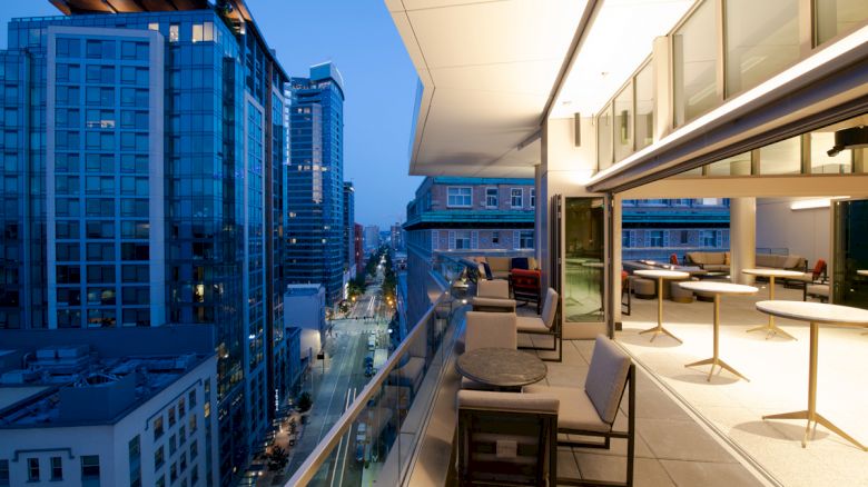 A modern outdoor terrace with seating overlooks a cityscape of tall buildings and streets at dusk, creating an inviting urban atmosphere.