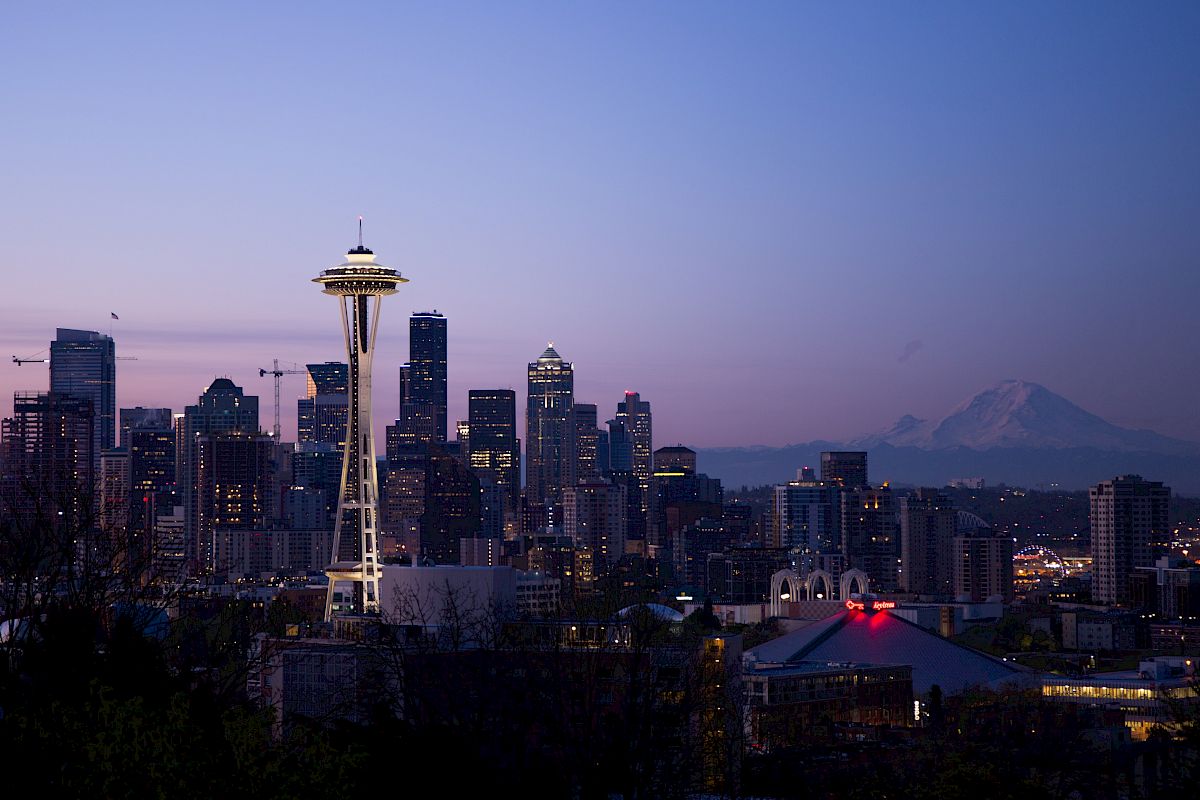 The image shows Seattle's skyline at dusk, featuring the Space Needle and Mount Rainier in the background, under a purple and blue sky.