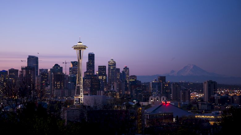The image shows Seattle's skyline at dusk, featuring the Space Needle and Mount Rainier in the background, under a purple and blue sky.