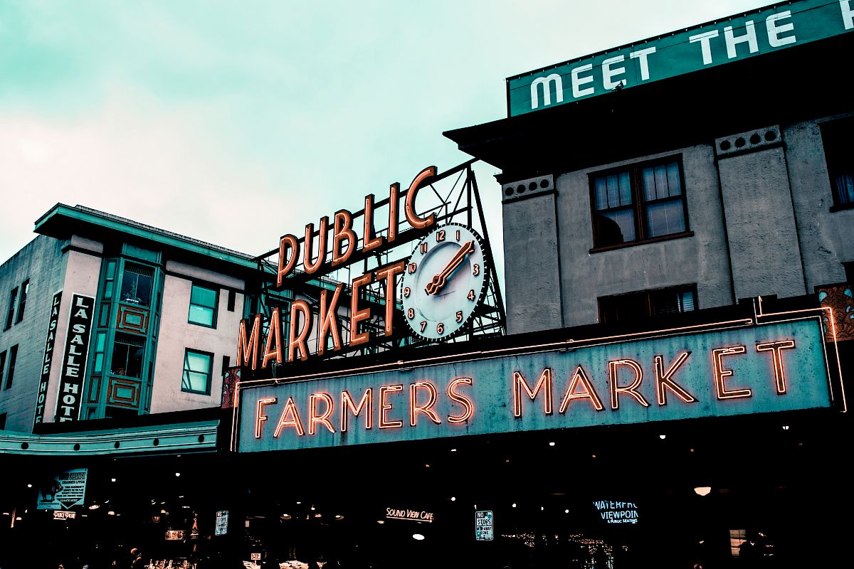 The image shows a neon sign reading "Public Market Center" and "Farmers Market" with a clock above, set against an urban building backdrop.