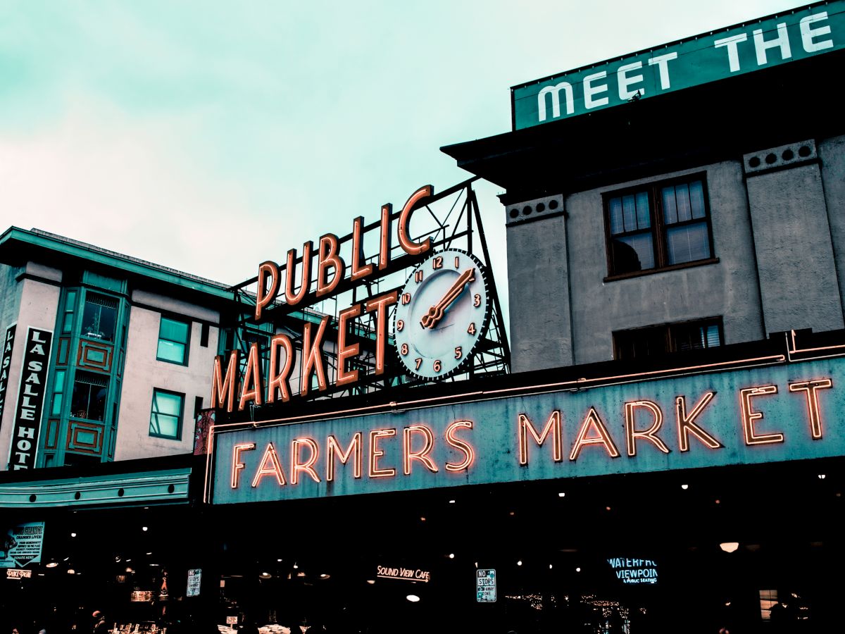 The image shows a neon sign reading "Public Market Center" and "Farmers Market" with a clock above, set against an urban building backdrop.