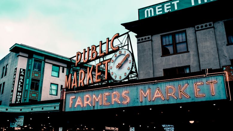 The image shows a neon sign reading "Public Market Center" and "Farmers Market" with a clock above, set against an urban building backdrop.