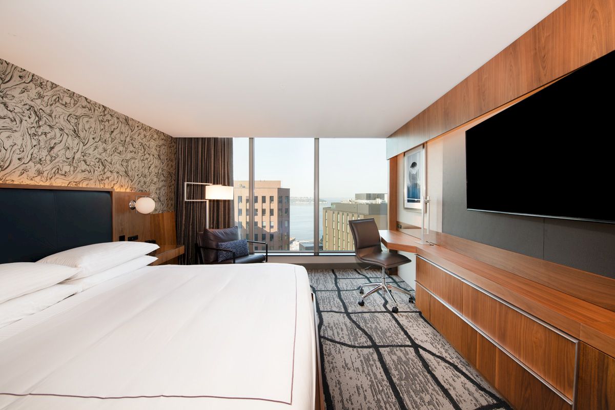 A modern hotel room with a large bed, desk, chair, and wall-mounted TV, featuring a view of the cityscape through a large window.
