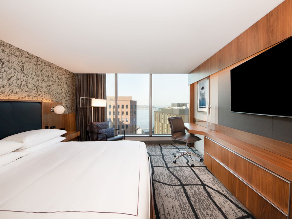 A modern hotel room with a large bed, desk, chair, and wall-mounted TV, featuring a view of the cityscape through a large window.