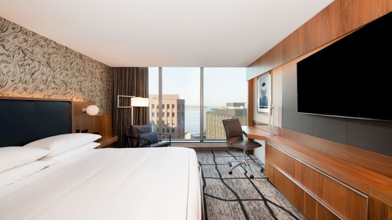 A modern hotel room with a large bed, desk, chair, and wall-mounted TV, featuring a view of the cityscape through a large window.
