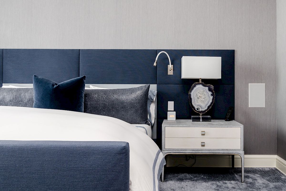 A modern bedroom with a blue and white color scheme, featuring a bed, pillows, and a nightstand with a lamp and decorative items.
