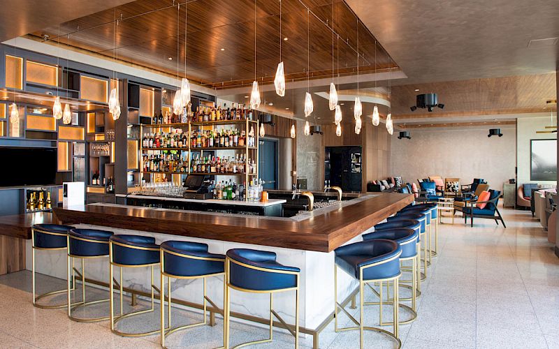 A modern bar with wooden accents, hanging lights, and plush seating. The bar is stocked with various bottles, surrounded by blue stools.