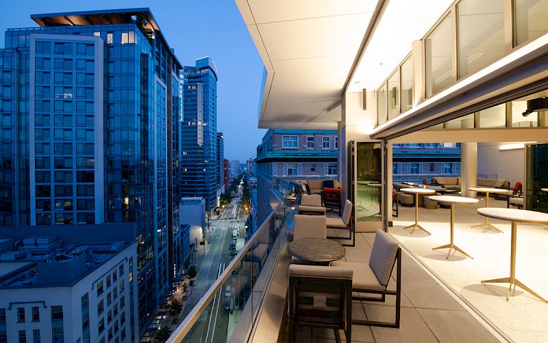 A modern rooftop terrace with seating areas overlooks a cityscape of tall buildings during dusk, featuring glass windows and urban ambiance.