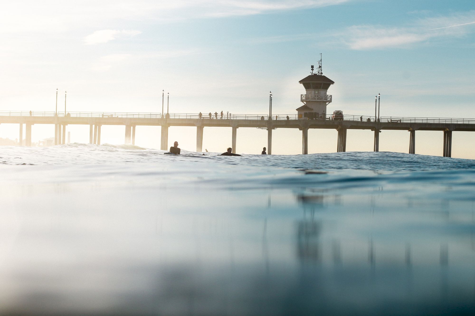 A serene view of a pier extending over the ocean with a watchtower, waves, and silhouettes of people, under a clear sky.